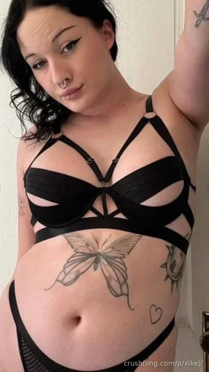 Woman with dark hair and tattoos wears black strappy lingerie in indoor setting.