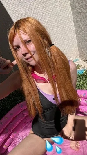 Woman with long red hair in a black and purple swimsuit sitting on a pink inflatable pool outdoors.