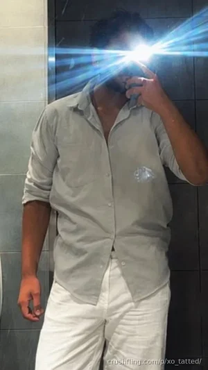 Man in light gray button-up shirt and white pants standing against tiled wall, holding phone to