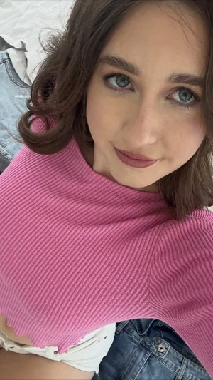 Woman with brown hair and blue eyes wearing pink top and denim shorts, lying on bed.