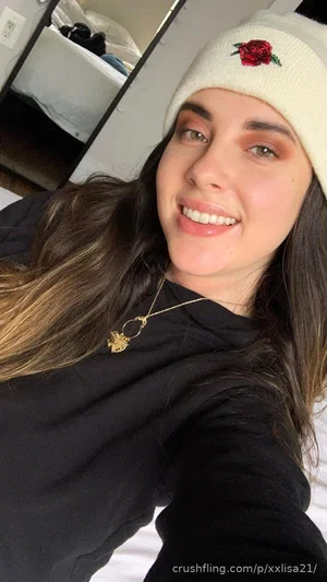 Woman with long brown hair wearing black top and white beanie with red rose, smiling in bedroom