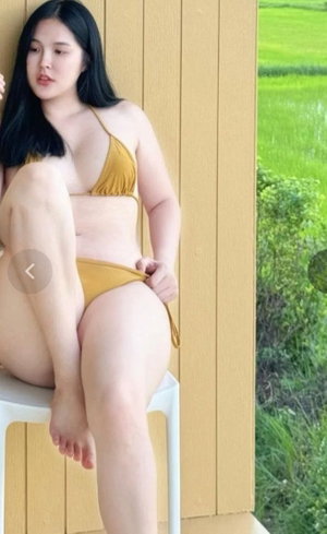 Woman in yellow bikini sitting on stool near wooden wall, green field in background.