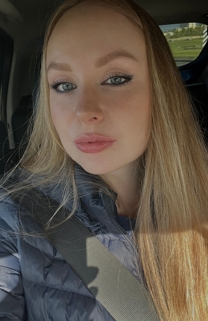Woman with blonde hair and blue eyes wearing a puffer jacket in a car.