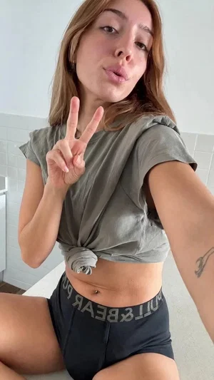 Woman in olive crop top and black shorts posing indoors, making peace sign.