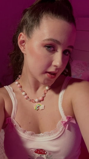 Woman in pink lace tank top with rainbow pendant necklace, pink lighting, indoor setting.