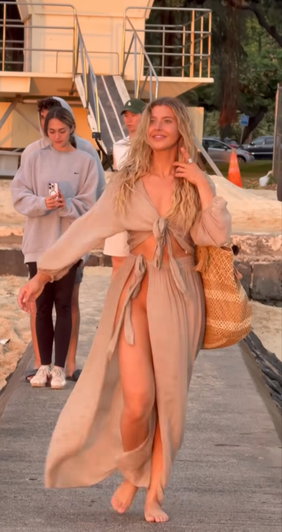 Blonde woman in beige outfit walking barefoot on beach path.