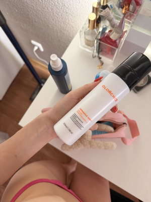 Woman in pink underwear holding a white bottle, sitting at a vanity with beauty products.