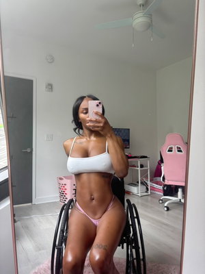 Woman in white top and pink underwear in wheelchair, taking mirror selfie in bedroom.