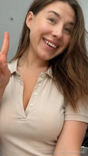 Woman with long brown hair in a beige polo shirt, smiling and waving indoors.