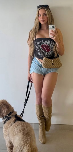 Woman with long blonde hair in a tank top and denim shorts, holding a phone and dog leash, standing