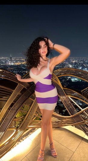 Woman in purple and white dress posing on balcony overlooking city at night.