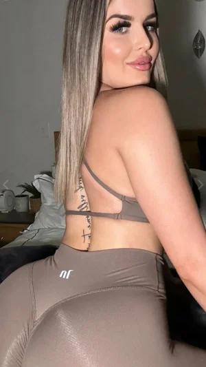 Woman with long blonde hair in gray sports bra and leggings, posing in bedroom.