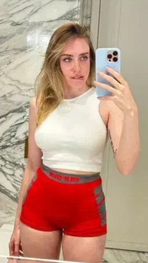 Woman with blonde hair in white crop top and red shorts taking mirror selfie in marble bathroom.