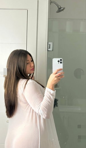 Long-haired woman in sheer top takes bathroom mirror selfie.