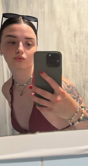 Woman in maroon top with tattoos takes mirror selfie indoors.