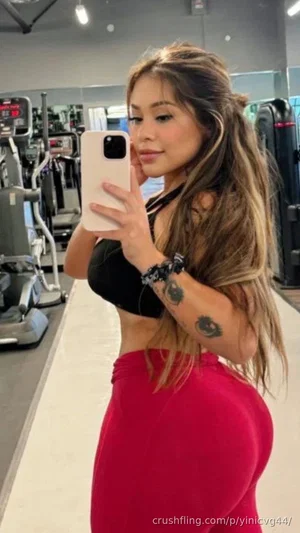 Woman with long hair in black crop top and red skirt taking mirror selfie in gym.