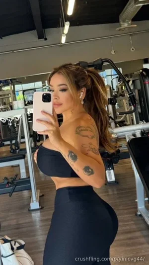 Woman with tattoos in black workout clothes taking mirror selfie in gym.