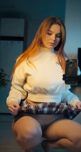 Red-haired woman in white sweatshirt and plaid skirt taking selfie indoors.