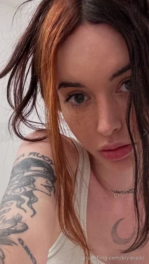Woman with tattoos on arm and chest, wearing beige tank top, indoors.