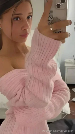 Woman in pink off-shoulder ribbed sweater taking mirror selfie in bedroom.
