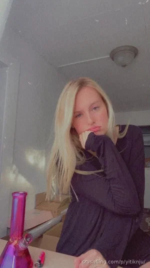 Blonde woman in dark long-sleeve top sitting indoors, hand near face, purple bong on table.