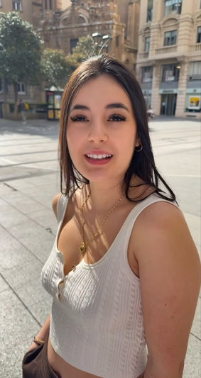 Woman with dark hair in white ribbed crop top, smiling in city plaza.