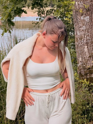 Woman in white crop top and pants outdoors near water and tree.