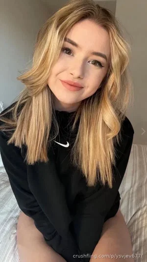 Woman with blonde hair wearing black hoodie, sitting on bed indoors.