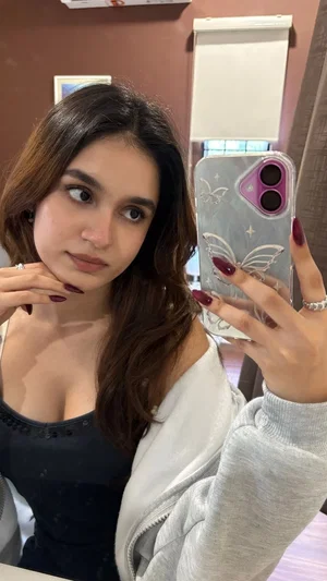 Woman with long brown hair in a black top and gray hoodie, taking mirror selfie indoors.