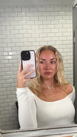 Blonde woman in white off-shoulder top taking mirror selfie in tiled bathroom.