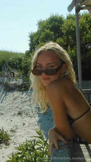 Blonde woman in bikini top on beach, wearing sunglasses, smiling at camera.
