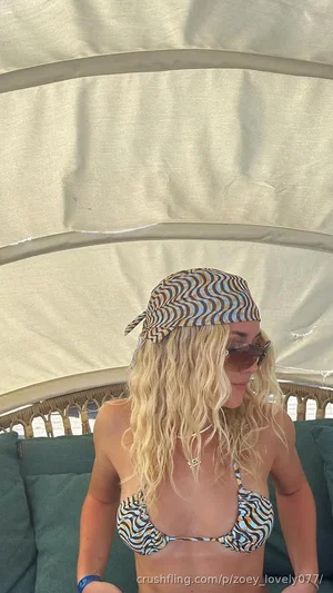 Blonde woman in patterned bikini top under canopy, wearing headscarf and sunglasses.