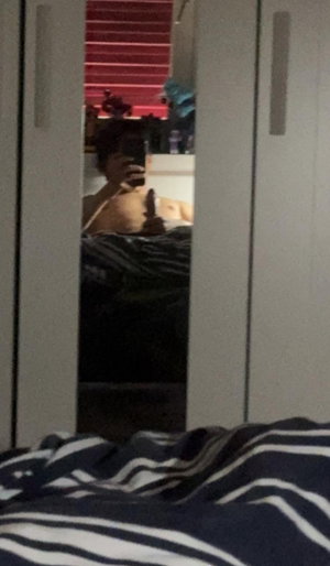 Woman with curly hair taking mirror selfie in bedroom, topless, striped bedding visible.
